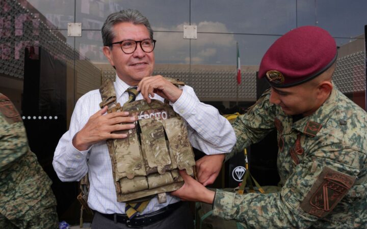 Ricardo Monreal ajusta un chaleco táctico con ayuda de un militar durante una exhibición.